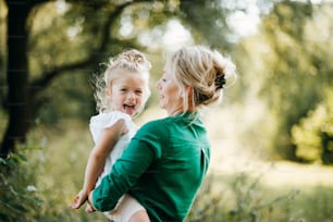 Beautiful young mother in green sunny summer nature holding her cute small daughter in the arms, having fun. Copy space.