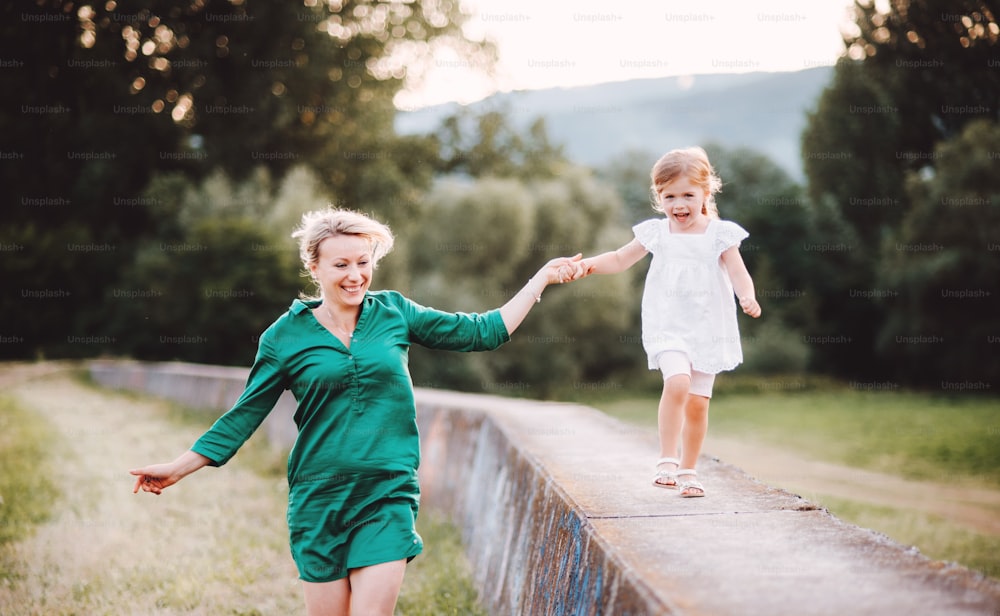 Young mother in nature with small daughter, having fun. A girl running on a concrete wall, holding hands.