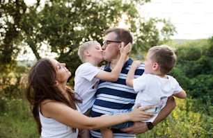 A cheerful young family holding two small sons outdoors in nature on a summer day, standing.