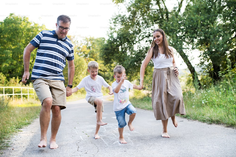 A young family with two small cheerful sons playing hopscotch barefoot on a road in park on a summer day, holding hands.