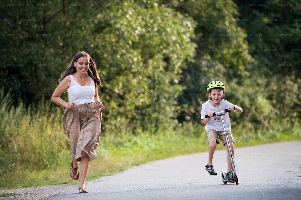 A cheerful small boy riding scooter and mother running on a road in park on a summer day.