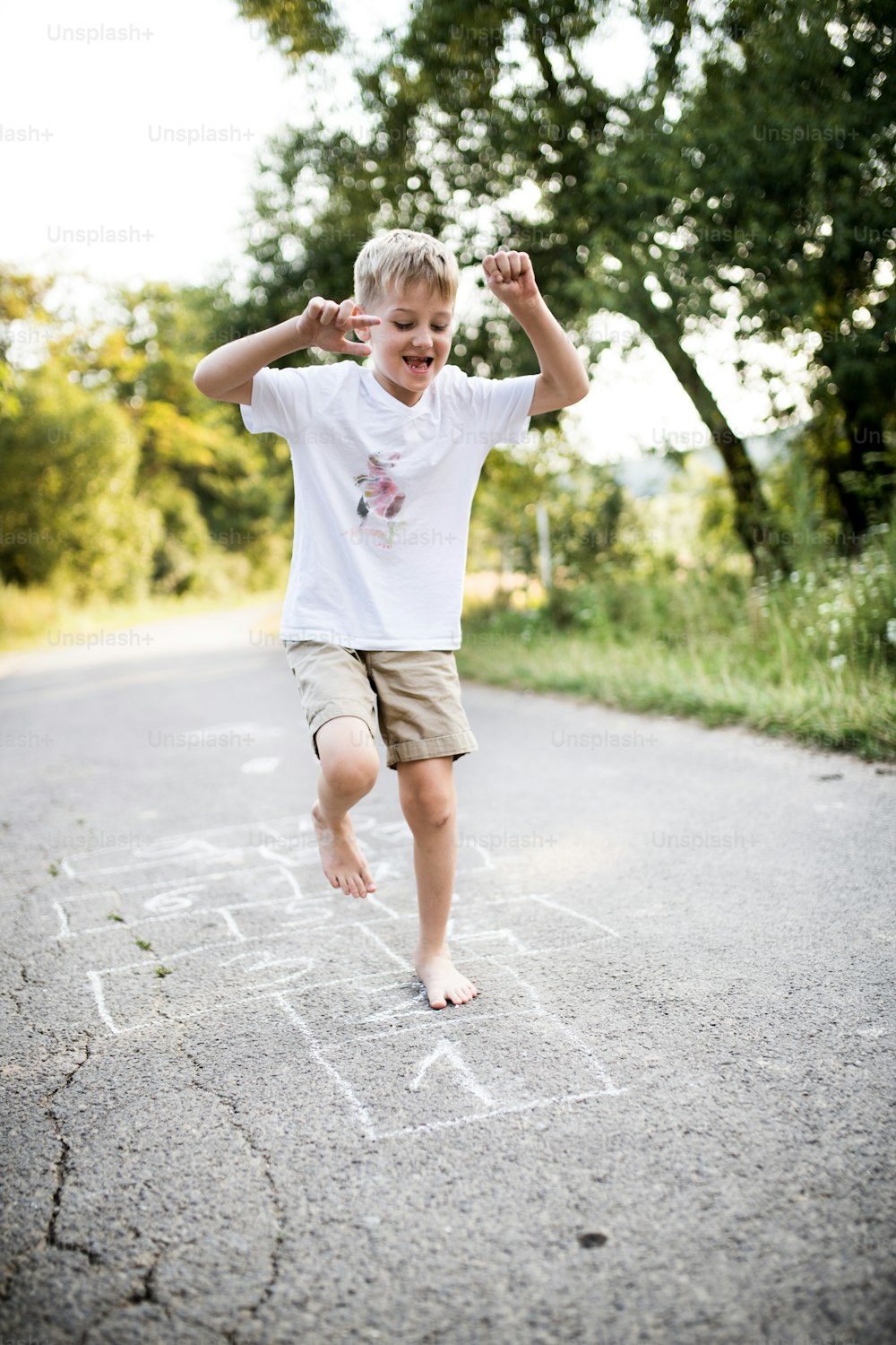 A barefoot cheerful small boy hopscotching on a road in park on a summer day.