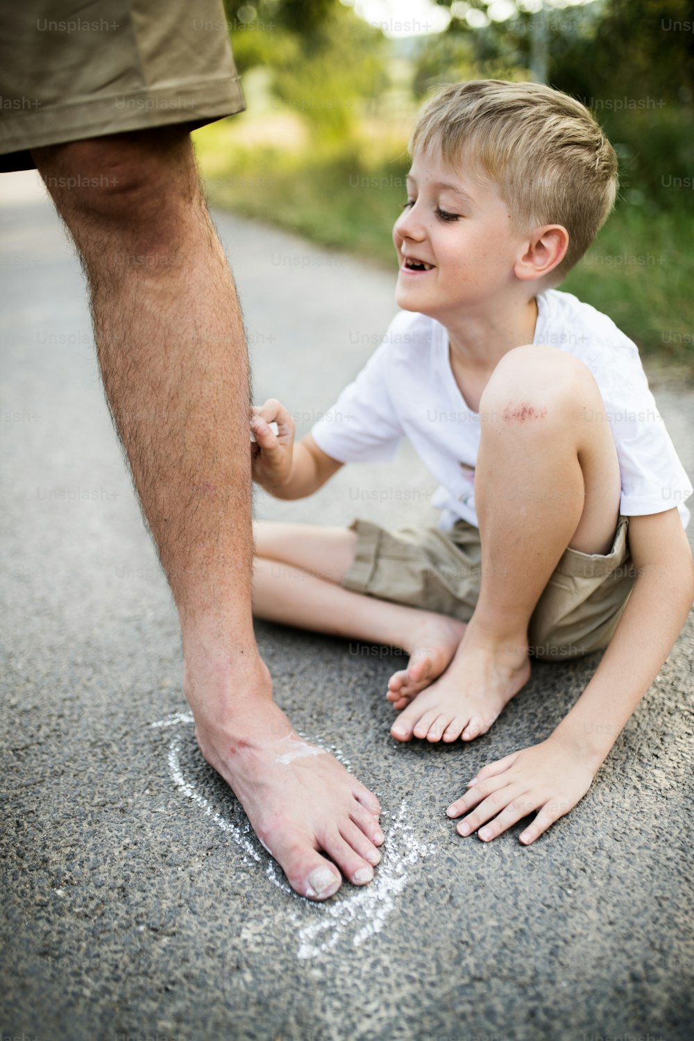 Ein kleiner Junge, der an einem Sommertag mit seinem Vater auf einer Straße im Park spielt und mit Kreide zeichnet.