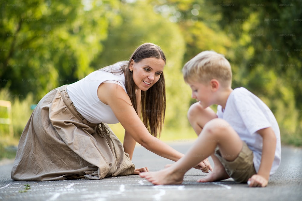 A small boy with mother drawing hopscotch with chalk on a road in park on a summer day.