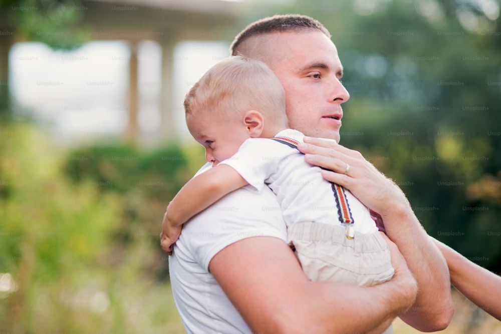 Young father holding a small crying toddler son in sunny summer nature, comforting him.