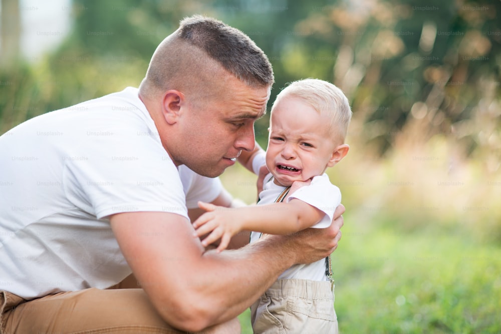 Young father holding a small crying toddler son in sunny summer nature, comforting him.