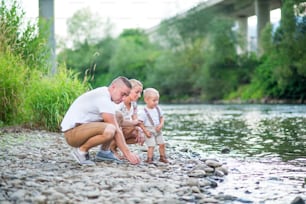 Família jovem com um menino pequeno que passa o tempo na natureza ensolarada do verão, brincando à beira do rio.