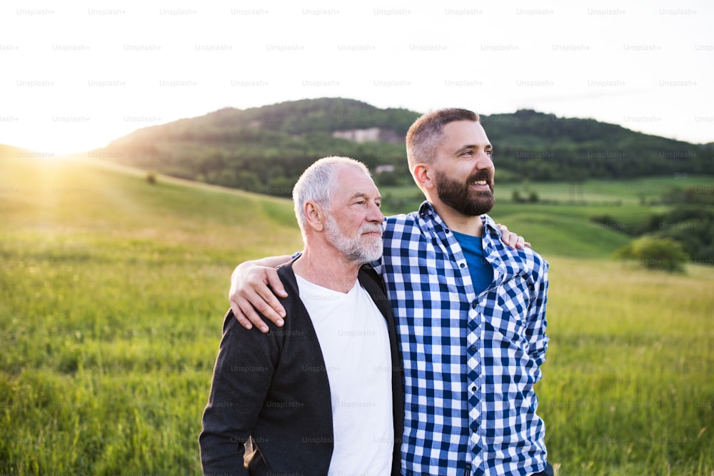 A portrait of a laughing adult hipster son with senior father in nature at sunset, arms around each other.