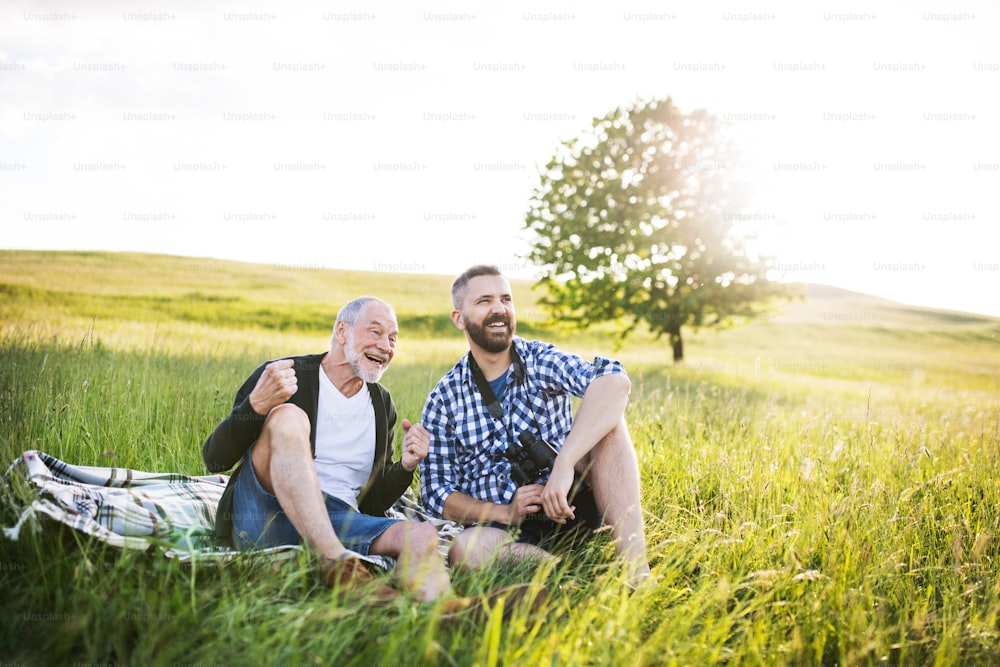 An adult hipster son with his senior father sitting on the grass at sunset in nature.