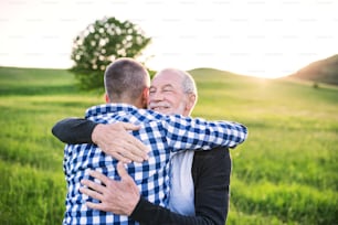 An adult hipster son with his senior father on a walk in nature at sunset, hugging. Copy space.