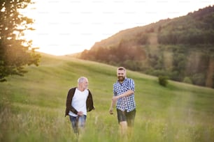 An adult hipster son with his senior father walking on a meadow in nature at sunset.