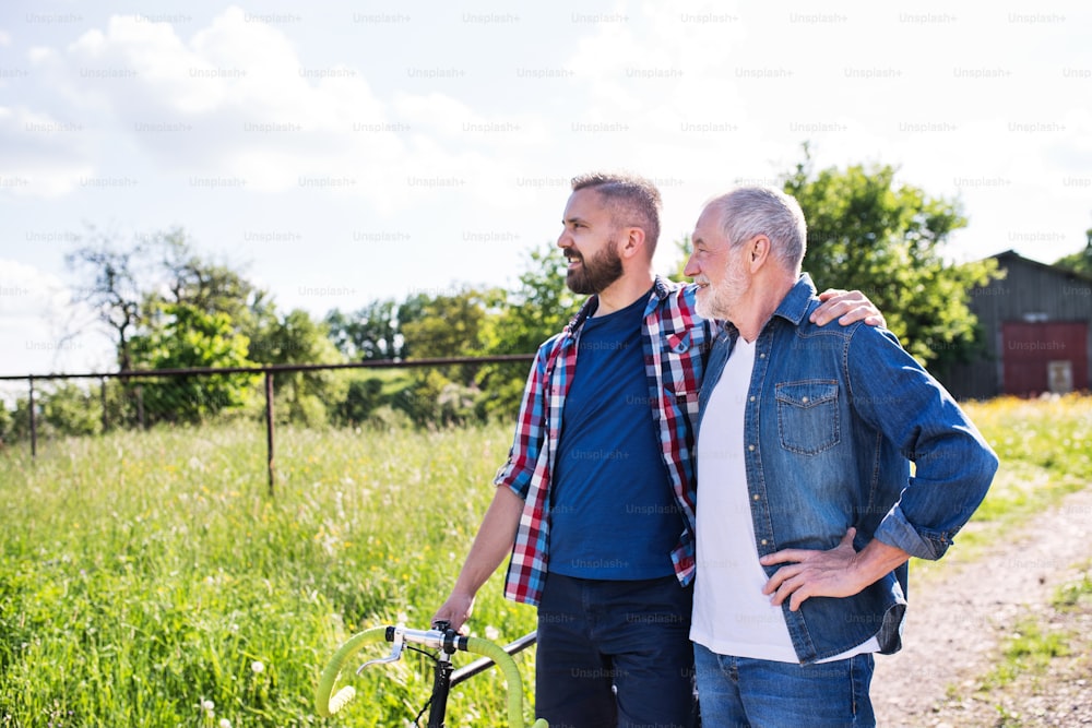 An adult hipster son with bicycle and senior father on a walk, arms around each other. Sunny nature. Copy space.