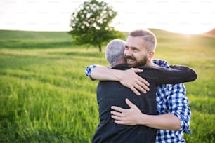 An adult hipster son with his senior father on a walk in nature at sunset, hugging. Copy space.