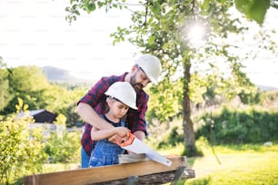 Father and a small daughter with helmets and a saw outside, cutting wood.