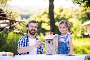Mature father with a small daughter outside, painting. Wooden birdhouse or bird feeder making.