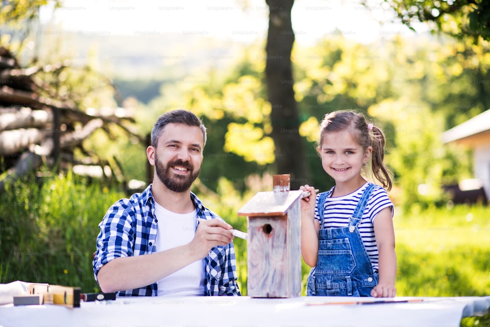 Mature father with a small daughter outside, painting. Wooden birdhouse or bird feeder making.