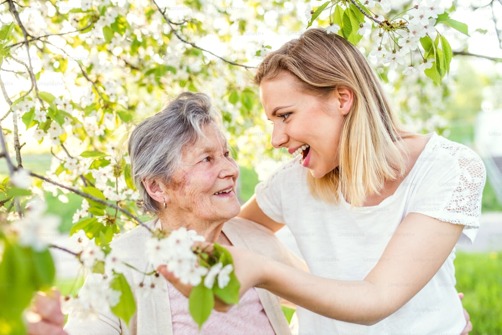 Ältere Großmutter und eine erwachsene Enkelin stehen unter einem Baum draußen in der Frühlingsnatur und lachen.
