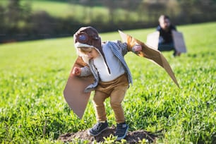 Happy toddler boy with hat, goggles and wings playing outside with unrecognizable father in spring nature. Pilot and flying concept.