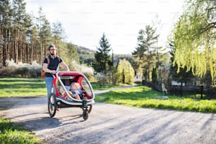 2人の幼児を持つ父親が、春の自然の中で外を散歩するジョギング用ベビーカーに座っている。