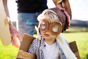 Niño feliz con sombrero, gafas y alas jugando afuera con un padre irreconocible en la naturaleza primaveral. Concepto de piloto y vuelo.