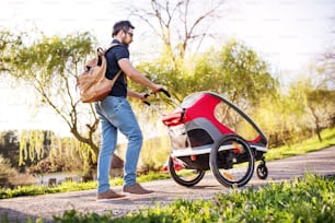 バックパックとジョギング用ベビーカーを持つ父親が、春の自然の中を外を散歩している。