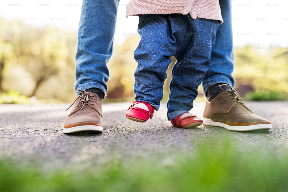 Primeros pasos de una niña pequeña afuera en la naturaleza primaveral. Padre irreconocible sosteniendo a su hija al caminar.