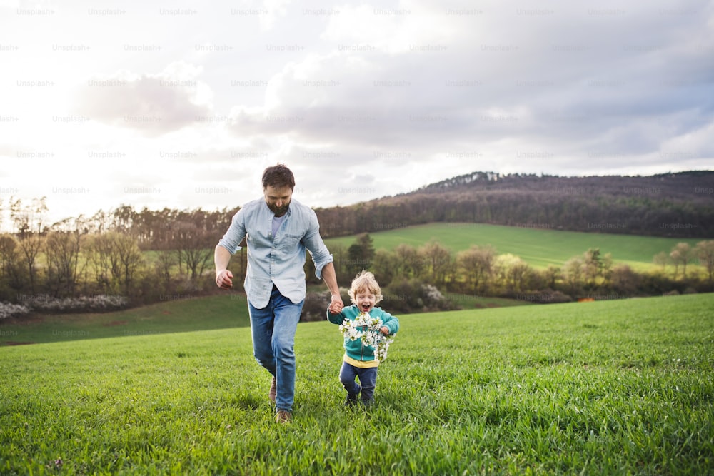 Un padre con su hijo pequeño corriendo afuera en la naturaleza verde y soleada de primavera, tomados de la mano.