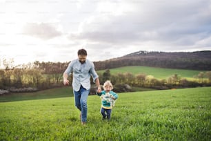 Un padre con su hijo pequeño corriendo afuera en la naturaleza verde y soleada de primavera, tomados de la mano.