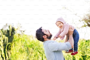 A handsome father holding his toddler daughter outside in green sunny spring nature.