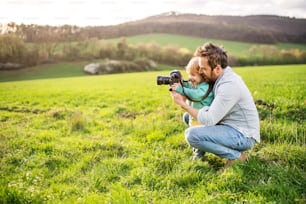 Ein Vater und sein kleiner Sohn mit einer Kamera draußen in der grünen, sonnigen Frühlingsnatur.