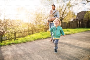 Ein Vater mit seinen kleinen Kindern draußen bei einem sonnigen Frühlingsspaziergang. Ein Mädchen in einer Babytrage und ein Junge rennt.