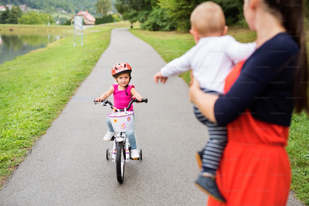 Linda niña feliz con su madre y su hermano menor en la naturaleza junto al lago. La chica está montando en bicicleta y sonriendo. Cámara lenta.