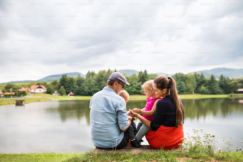 Família jovem e feliz passando tempo juntos do lado de fora na natureza verde, sentados em uma margem do lago. Vista traseira.