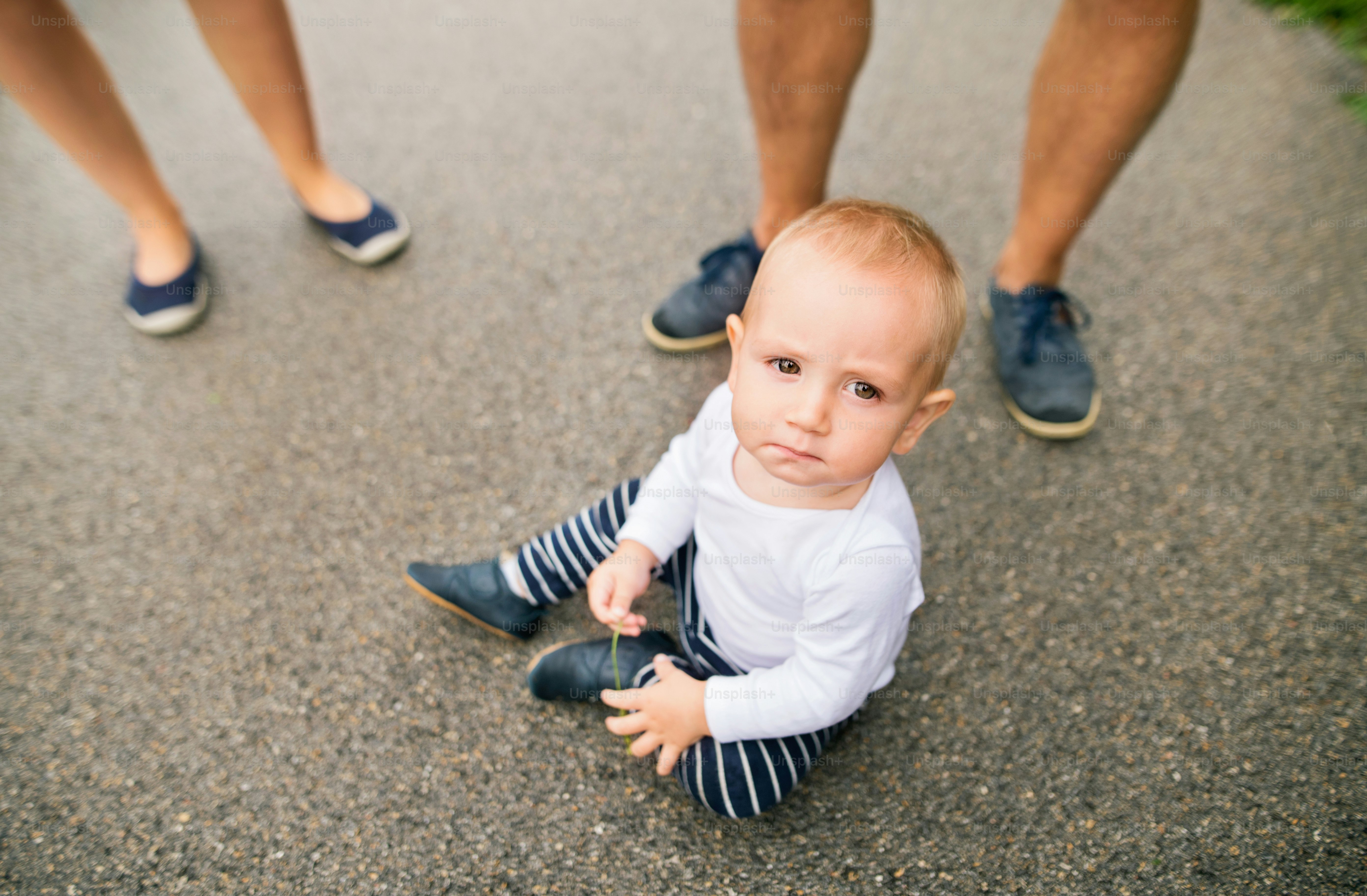 Little boy with unrecognizable parents sitting on the pavement in nature. Summer time