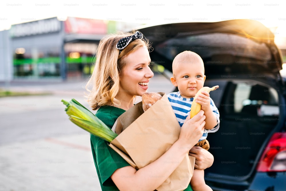 Schöne junge Mutter mit ihrem kleinen kleinen Sohn vor einem Supermarkt, der eine Einkaufstasche aus Papier hält. Frau mit einem Jungen, der am Auto steht.
