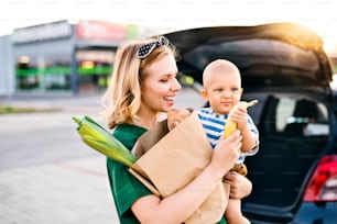 Beautiful young mother with her little baby son in front of a supermarket, holding paper shopping bag. Woman with a boy standing by the car.