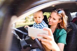 幼い息子を車に乗せた若い母親。運転しているふりをしている女性と男の子。スマートフォンで写真を撮る女性。