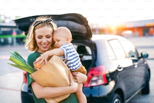 Beautiful young mother with her little baby son in front of a supermarket, holding paper shopping bag. Woman with a boy standing by the car.