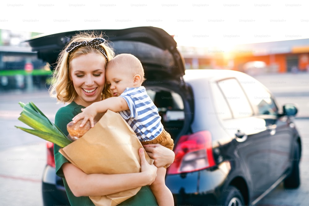 Hermosa madre joven con su pequeño hijo bebé frente a un supermercado, sosteniendo una bolsa de compras de papel. Mujer con un niño de pie junto al coche.