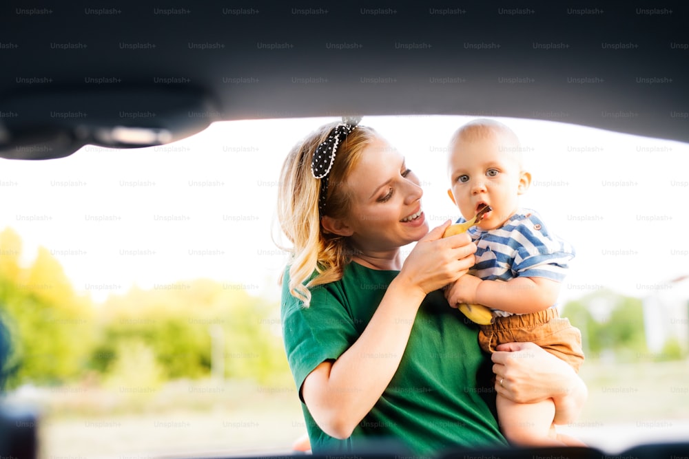 Jovem mãe segurando seu filhinho nos braços, de pé ao lado da parte de trás aberta do carro.