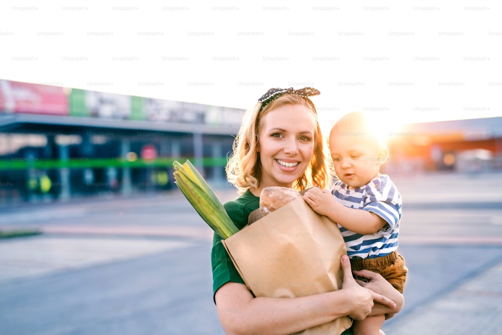 Hermosa madre joven con su pequeño bebé frente a un supermercado, sosteniendo una bolsa de compras de papel.