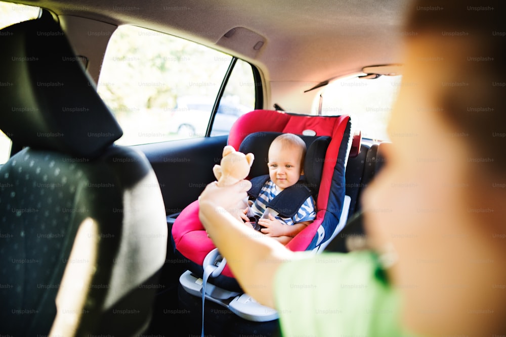 Junge Mutter mit ihrem kleinen Sohn im Auto. Kleiner Junge sitzt in einem Autositz, nicht wiederzuerkennende Frau, die ihm ein Spielzeug gibt.