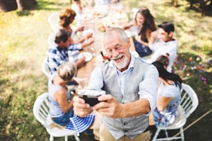 Family celebration outside in the backyard. Big garden party. A senior man taking selfie with a camera.