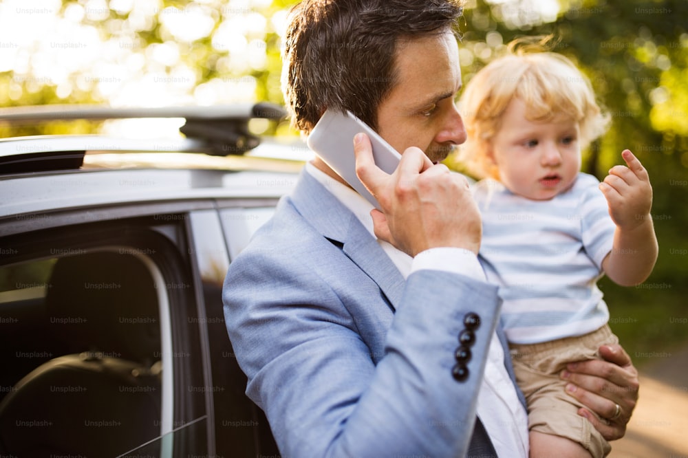 Young father holding his little boy in the arms by the car. Man with smartphone making a phone call.