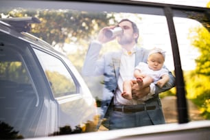 Padre joven cargando a su bebé. Hombre en el auto, tomando café. Disparado a través del vidrio.