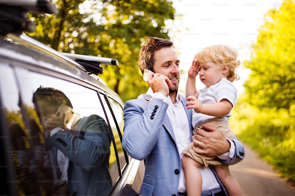 Der junge Vater hält seinen kleinen Jungen in den Armen und steigt ins Auto. Mann mit Smartphone telefoniert.