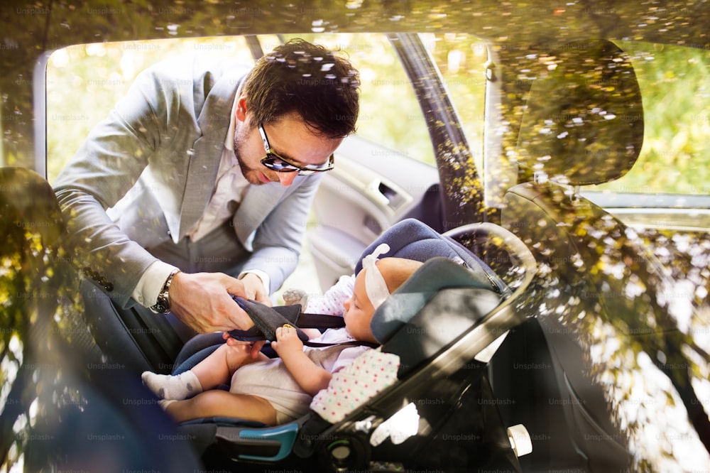 女の赤ちゃんを車に乗せるハンサムな若い男。シートベルトを締める父。ガラス越しに撃った。