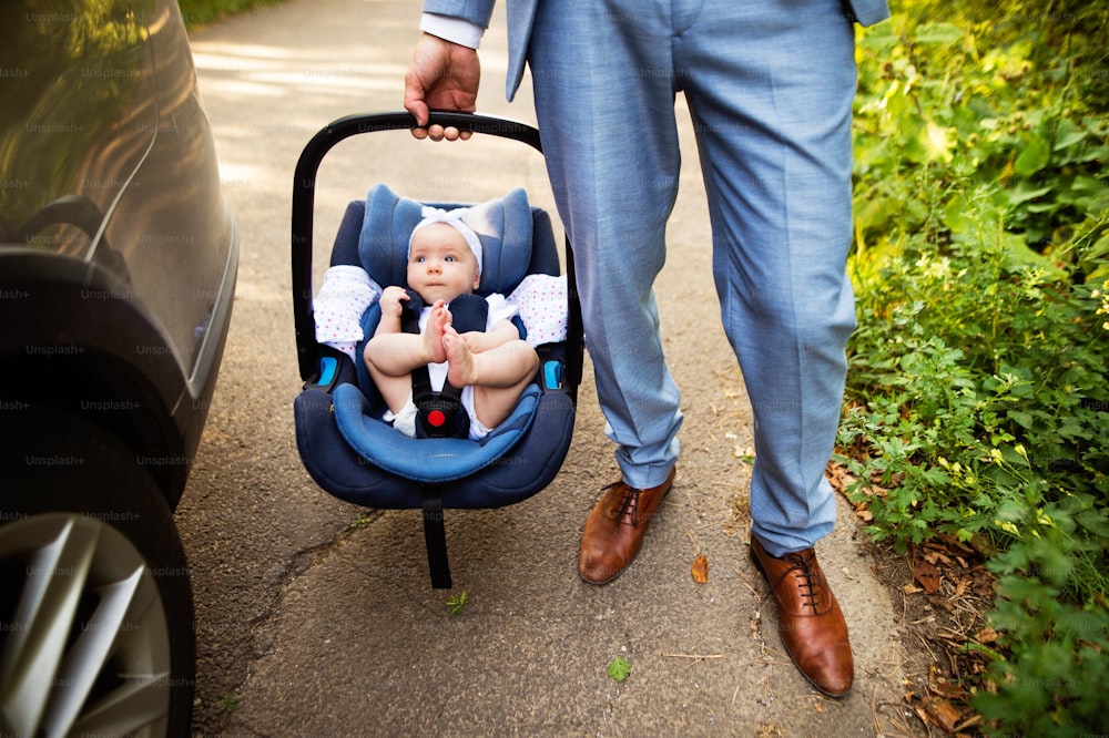 Homme méconnaissable portant sa petite fille dans un siège d’auto. Homme avec un bébé près de la voiture.