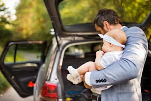 車のそばに立っている小さな女の赤ちゃんを抱く若い父親。ドアが開いた。