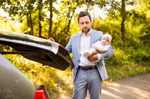 Padre joven sosteniendo a su pequeña bebé junto al auto. Hombre cerrando la puerta del maletero.